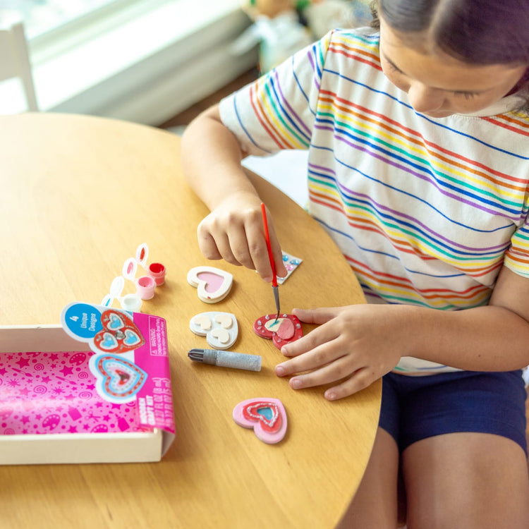 A kid playing with the Melissa & Doug Created by Me! Wooden Heart Magnets Craft Kit (4 Designs, 4 Paints, Stickers, Glitter Glue)