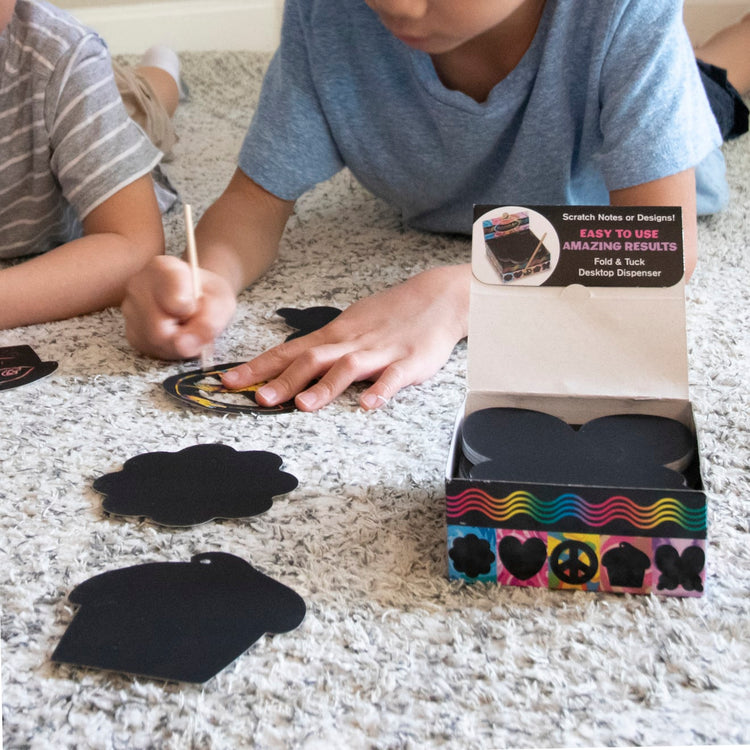 A kid playing with the Melissa & Doug Scratch Art® Box of 125 Friendship-Themed Shaped Notes in Desktop Dispenser (Approx. 3.5” x 3.5” Each Note)