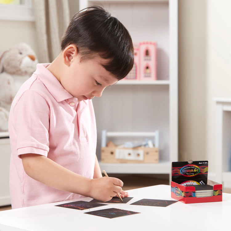 A kid playing with The Melissa & Doug Scratch Art Rainbow Mini Notes (125) With Wooden Stylus