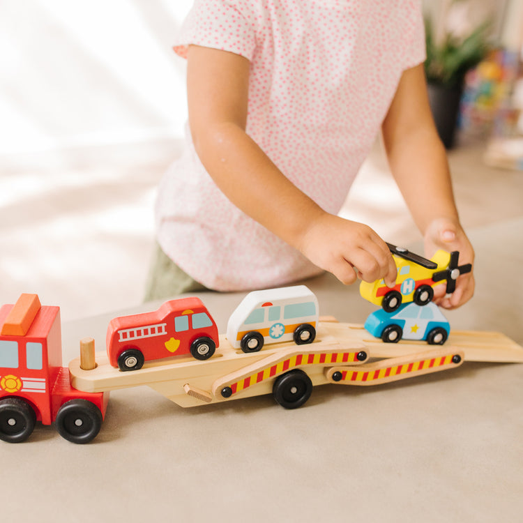 A kid playing with The Melissa & Doug Wooden Emergency Vehicle Carrier Truck With 1 Truck and 4 Rescue Vehicles