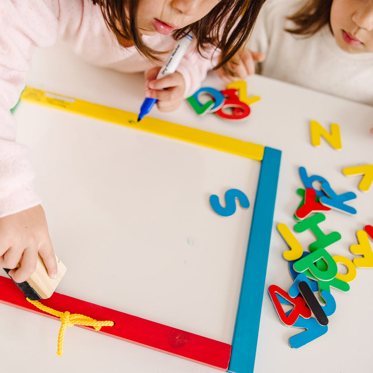 A kid playing with The Melissa & Doug Magnetic Chalkboard and Dry-Erase Board With 36 Magnets (Numbers and Uppercase Letters), Chalk, Eraser, and Dry-Erase Pen