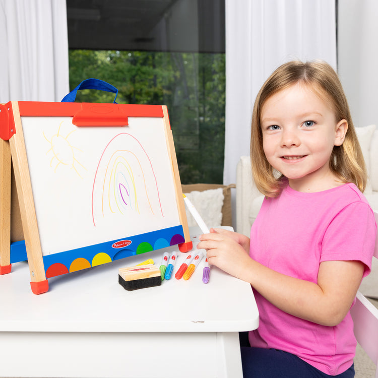 A kid playing with The Melissa & Doug Take-Along Double-Sided Wooden Tabletop Activity Art Easel; Portable Travel Toy with Whiteboard, Chalkboard, Arts and Crafts Accessories