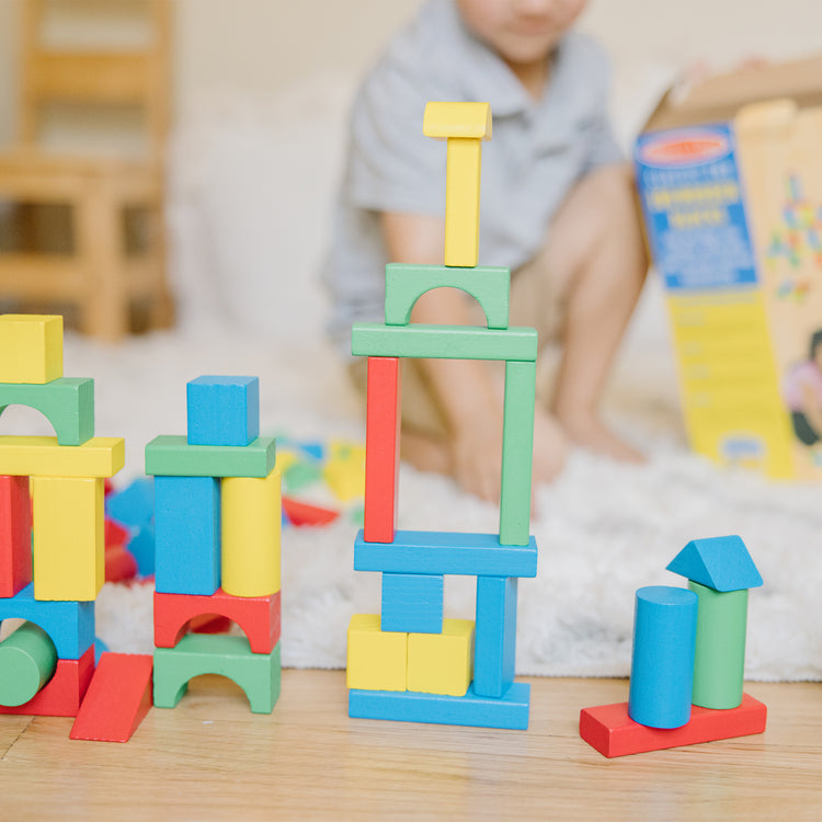 A kid playing with the Melissa & Doug Wooden Building Blocks Set - 100 Blocks in 4 Colors and 9 Shapes