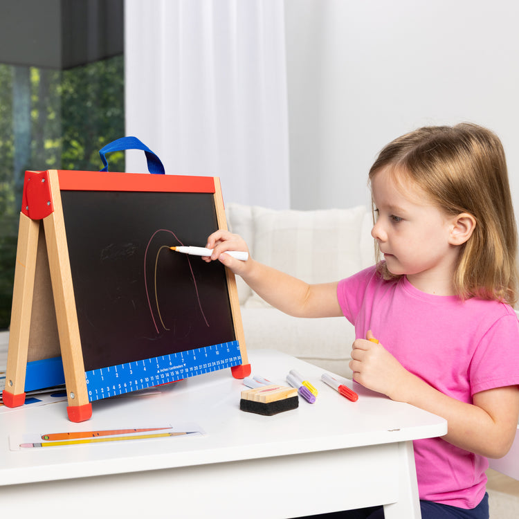 A kid playing with The Melissa & Doug Take-Along Double-Sided Wooden Tabletop Activity Art Easel; Portable Travel Toy with Whiteboard, Chalkboard, Arts and Crafts Accessories