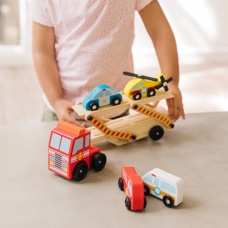 A kid playing with The Melissa & Doug Wooden Emergency Vehicle Carrier Truck With 1 Truck and 4 Rescue Vehicles