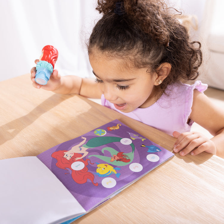 A kid playing with The Melissa & Doug Sticker Wow!™ 24-Page Activity Pad and Sticker Stamper, 300 Stickers, Arts and Crafts Fidget Toy Collectible Character – Disney Princess Ariel - for Kids Ages 3+