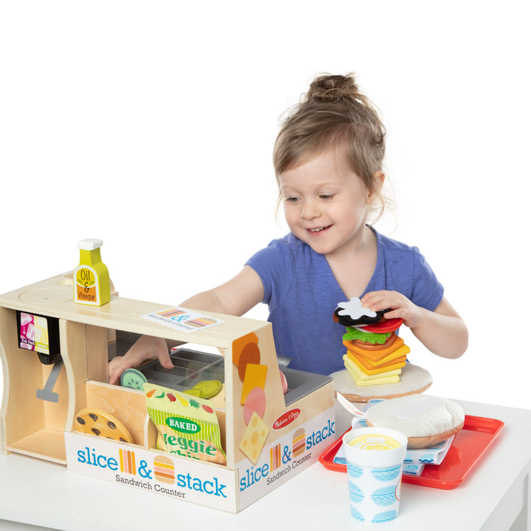 A child on white background with The Melissa & Doug Wooden Slice & Stack Sandwich Counter with Deli Slicer – 56-Piece Pretend Play Food Pieces