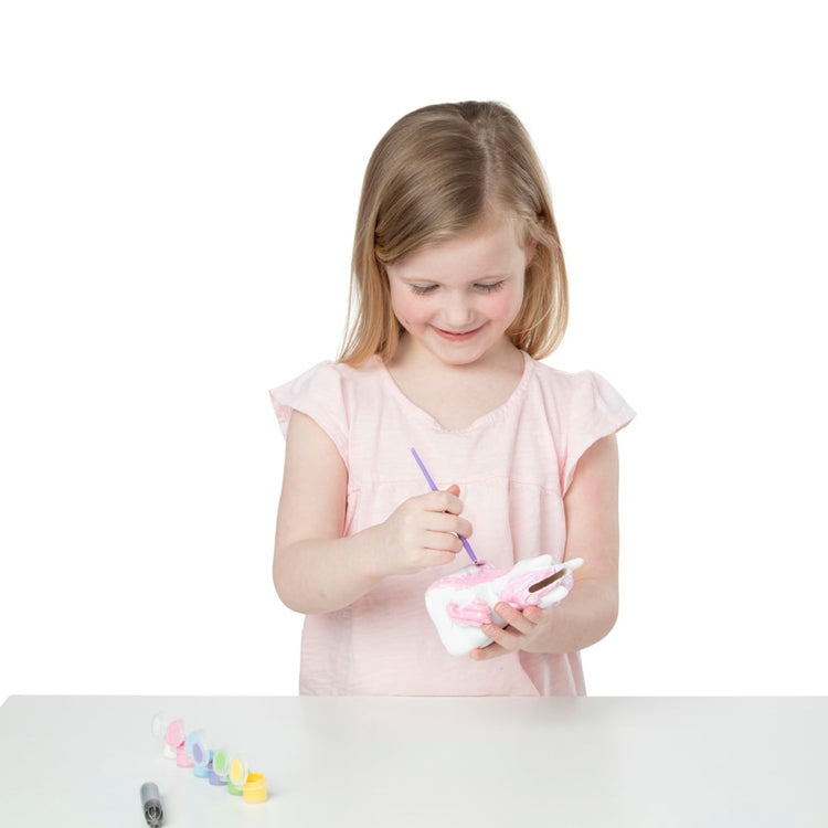 A child on white background with the Melissa & Doug Created by Me! Decorate-Your-Own Unicorn Bank Craft Kit With 6 Pots of Paint, Glitter Glue, Paintbrush