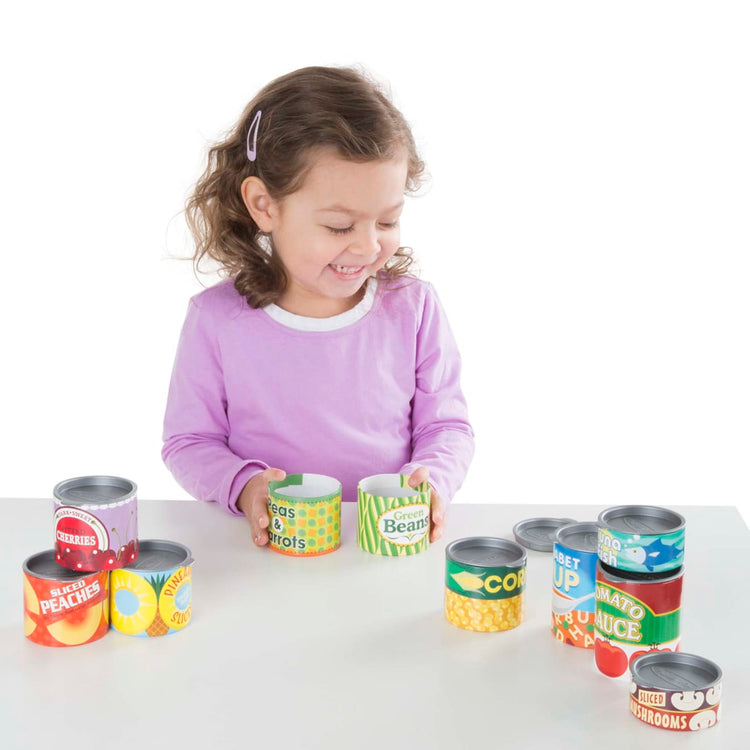 A child on white background with the Melissa & Doug Let's Play House! Grocery Cans Play Food Kitchen Accessory - 10 Stackable Cans With Removable Lids
