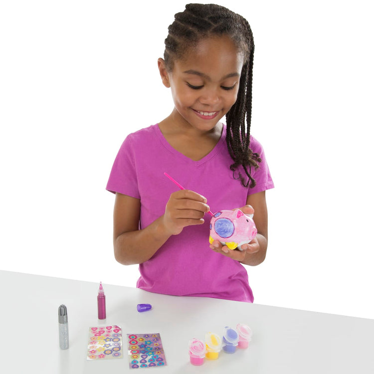 A child on white background with the Melissa & Doug Created by Me! Piggy Bank Craft Kit with 4 Pots of Paint, Brush, Glitter, Stickers