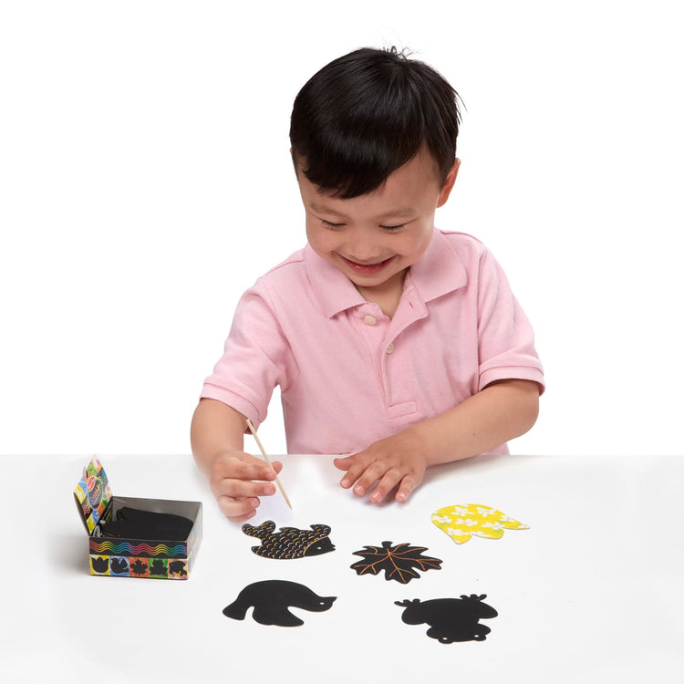 A child on white background with the Melissa & Doug Scratch Art® Box of 125 Nature-Themed Shaped Notes in Desktop Dispenser (Approx. 3.5” x 3.5” Each Note)